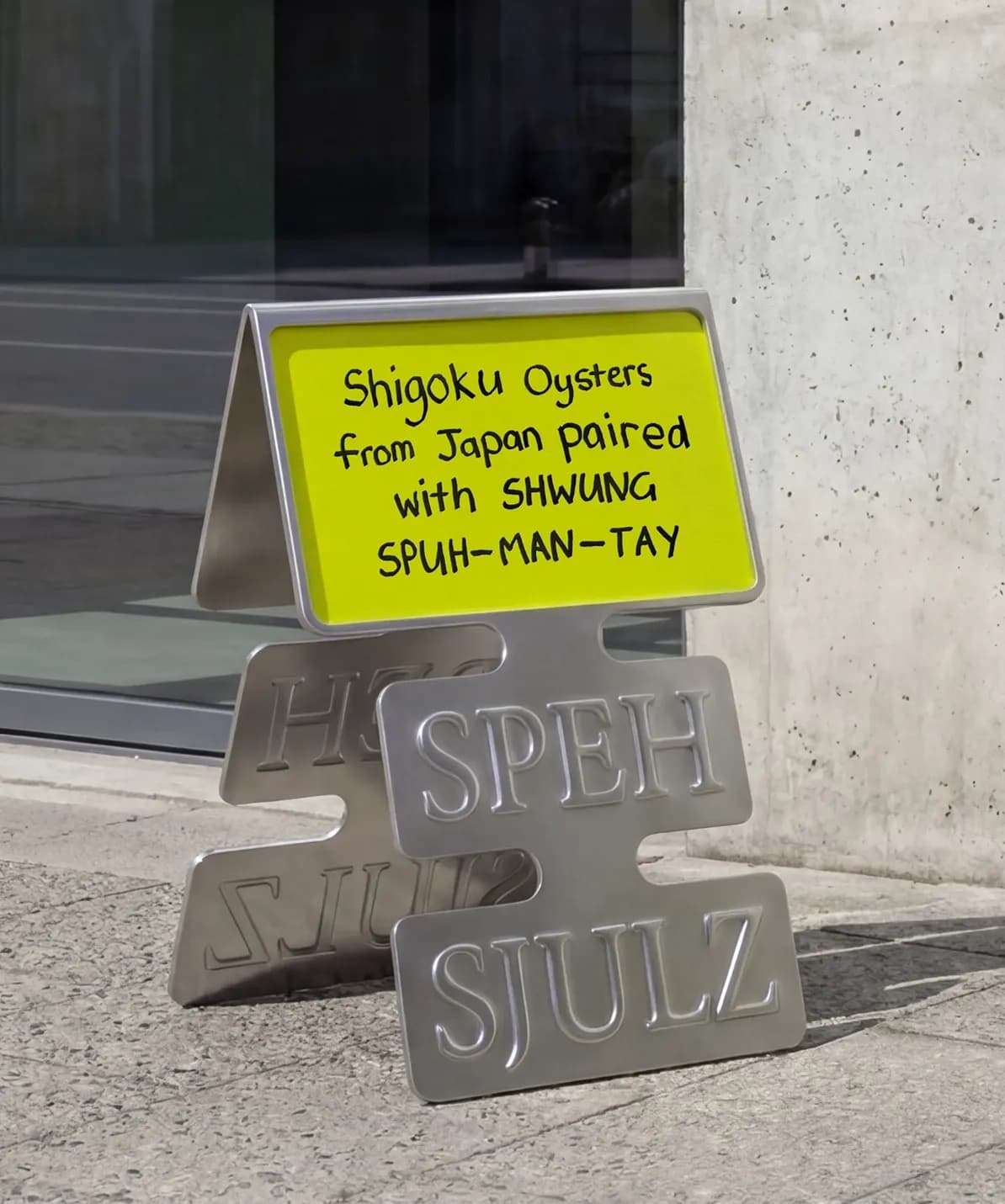 Shwung metal pavement sign in the die-cut label shape with "Speh Sjulz" embossed and a handwritten yellow-green card advertising Shigoku oysters from Japan paired with Shwung Spuh-man-tay, placed outside a restaurant entrance.