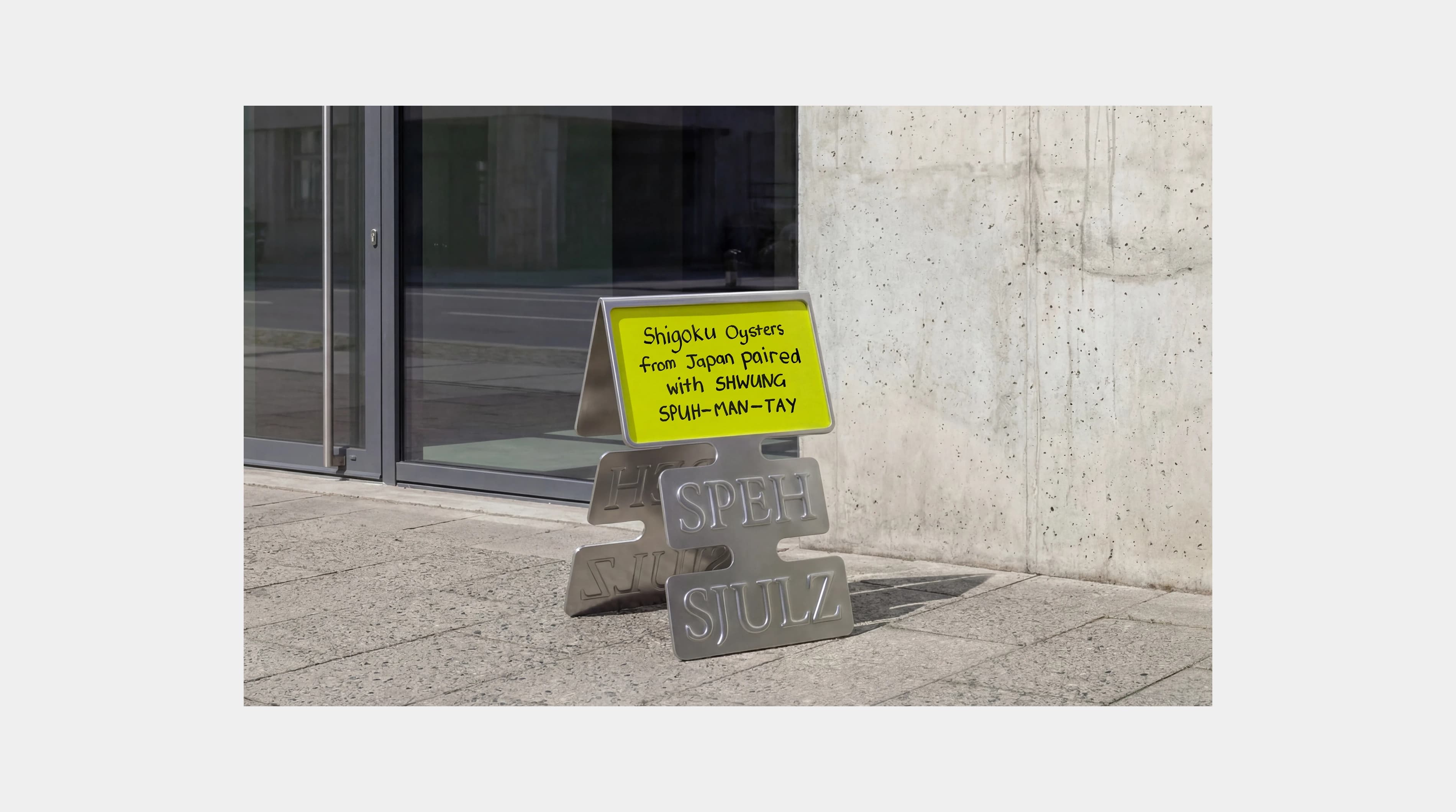 Shwung metal pavement sign in the die-cut label shape with "Speh Sjulz" embossed and a handwritten yellow-green card advertising Shigoku oysters from Japan paired with Shwung Spuh-man-tay, placed outside a restaurant entrance.