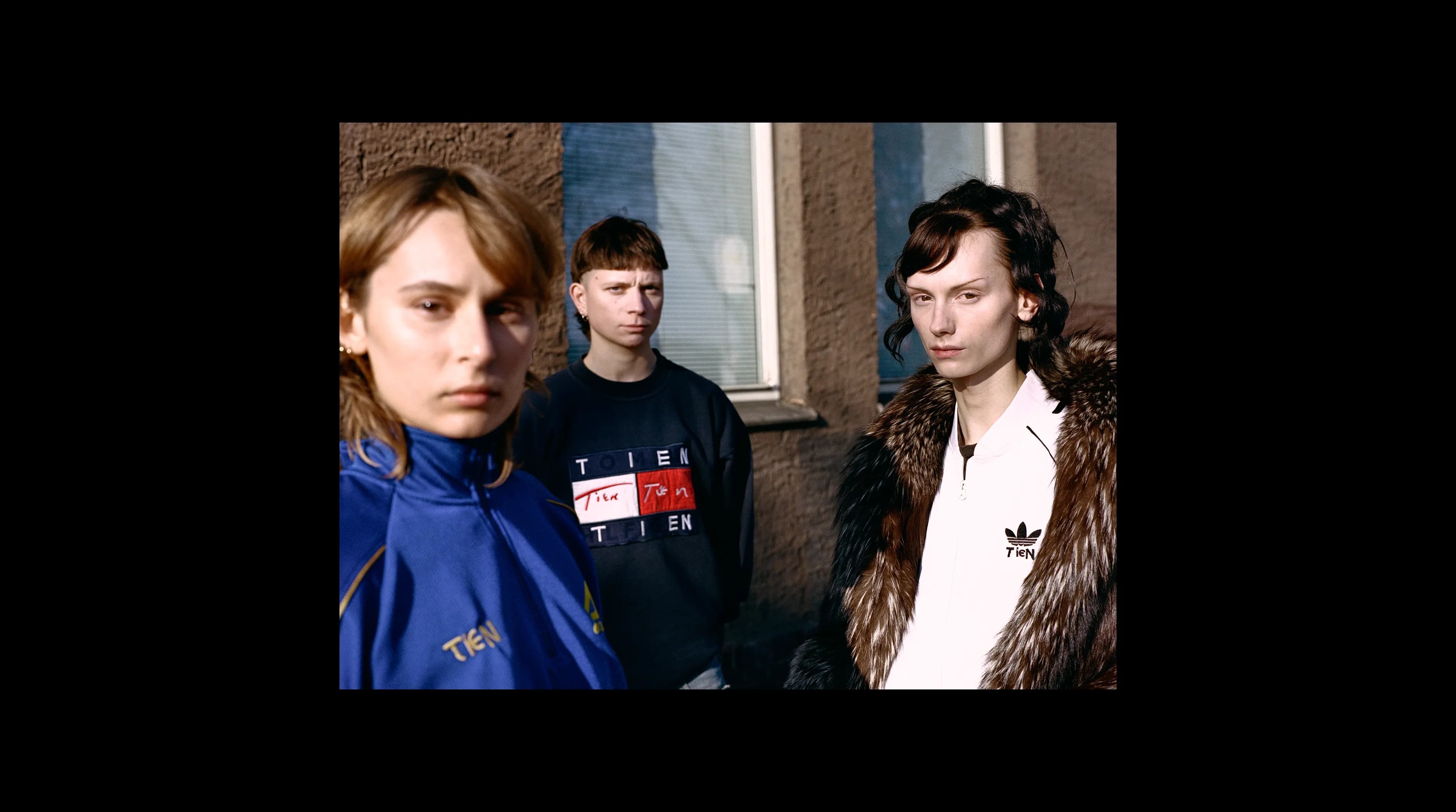 Three people wearing vintage sportswear reworked with the TIEN identity: an Adidas track jacket with TIEN embroidered on the chest, the Tommy Hilfiger sweatshirt with the flag logo replaced, and an Adidas zip-up with TIEN on the left chest.