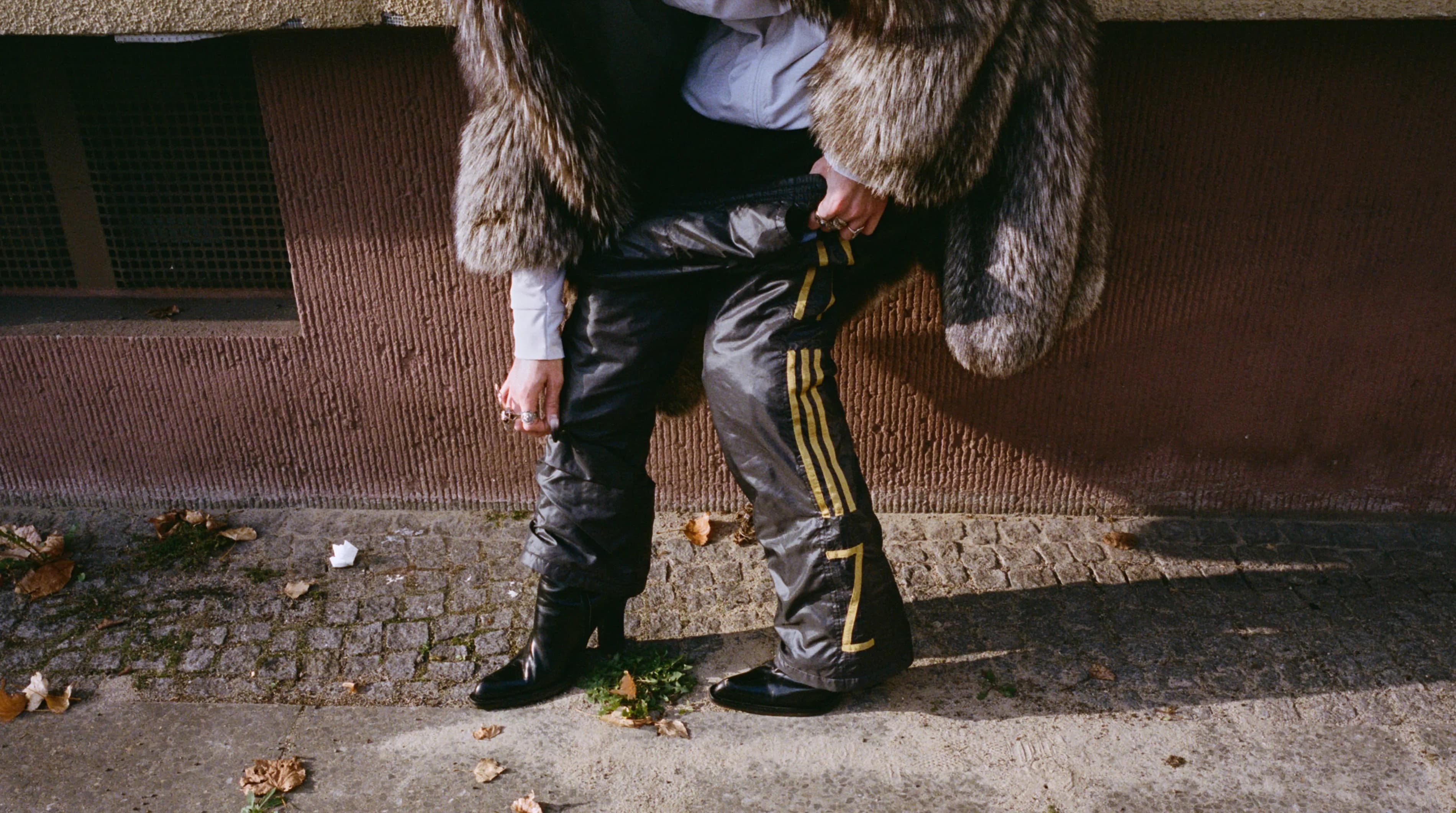 Close-up of a person standing on a cobblestone street wearing leather boots, a fur coat and black trackpants with yellow side stripes, shot for the TIEN campaign.