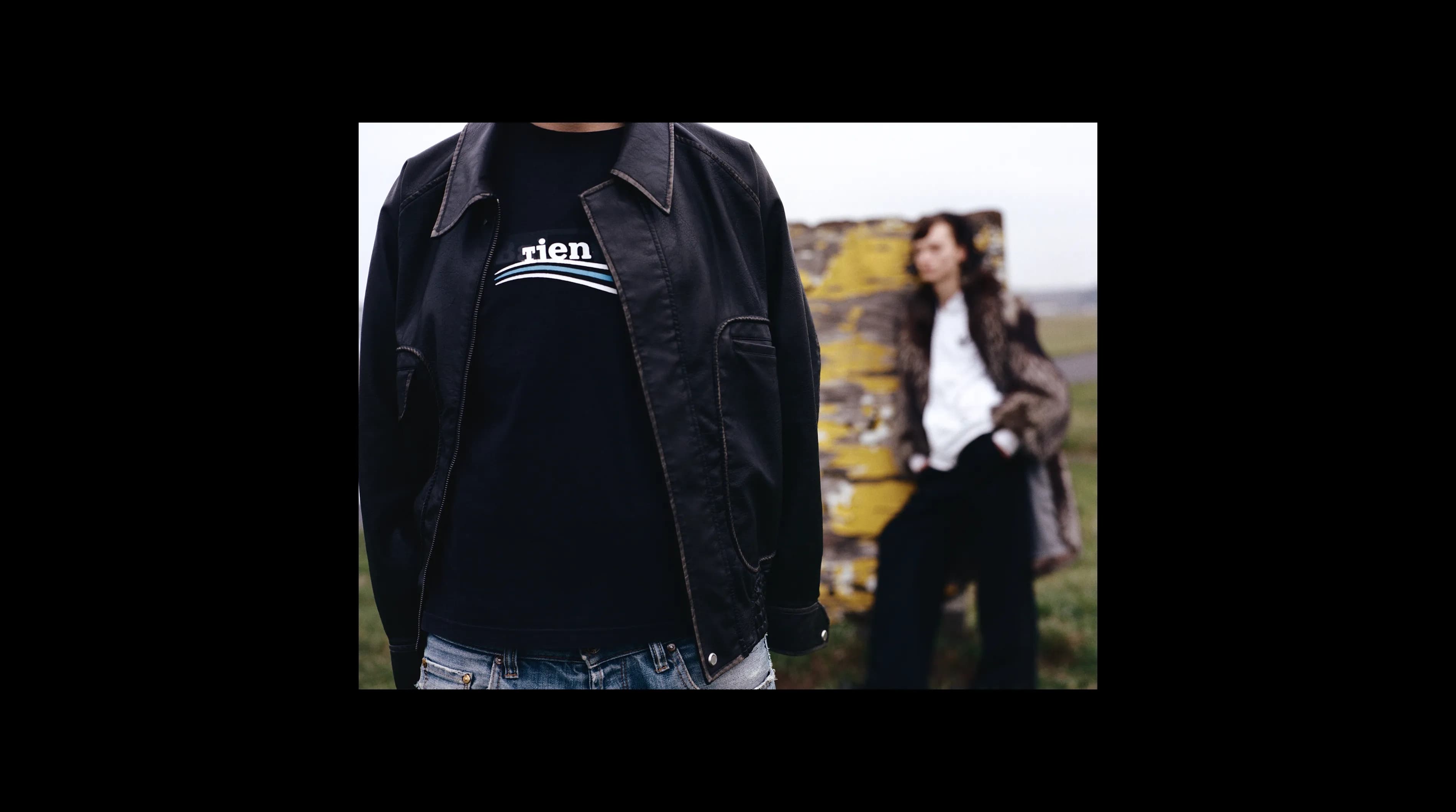 Person wearing a black t-shirt with a vintage Patagonia logo reworked to read "tien", layered under a black leather jacket, with a second person out of focus in the background.