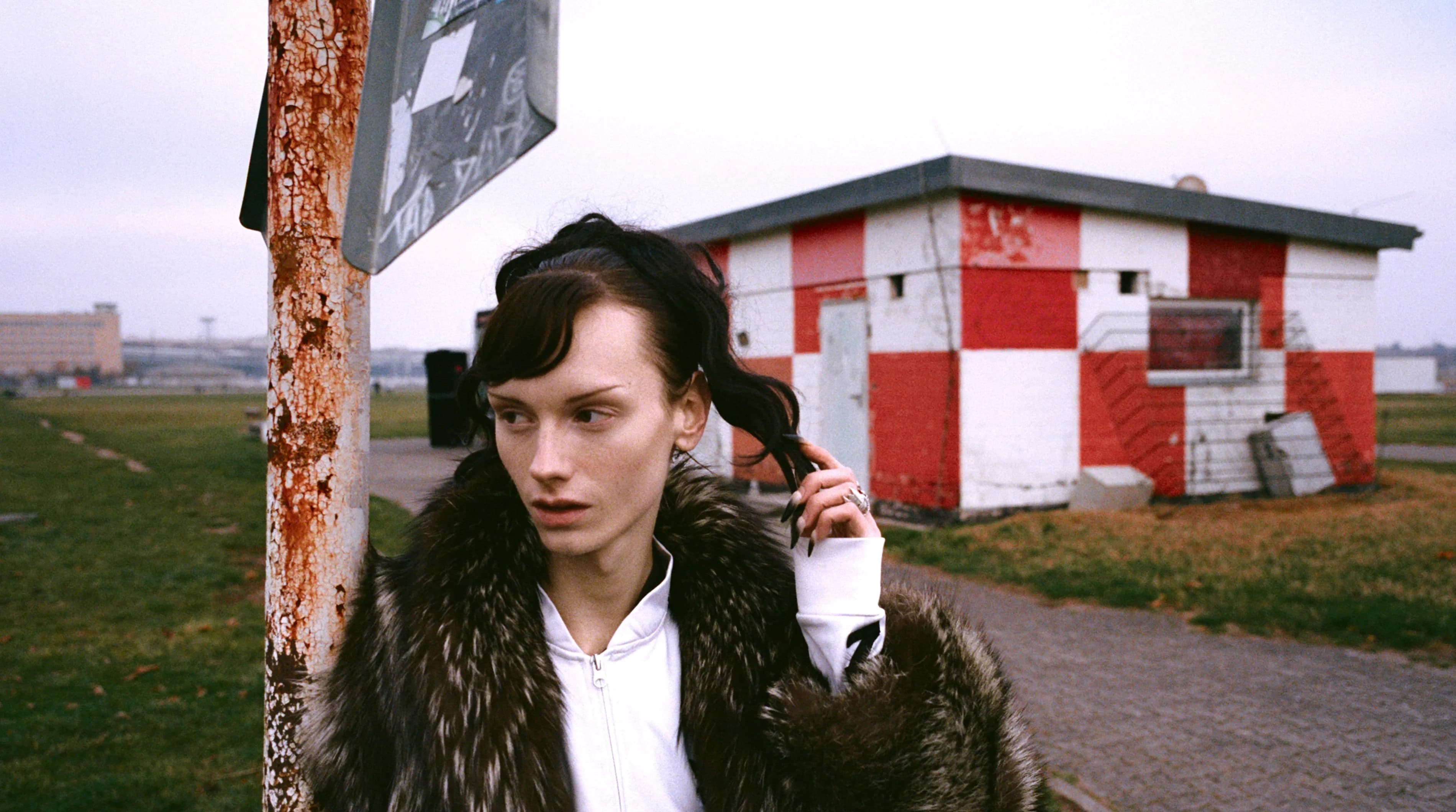 Person wearing a fur coat over a white Adidas zip-up with TIEN on the chest, standing next to a rusted street pole with a red and white checkered building in the background.