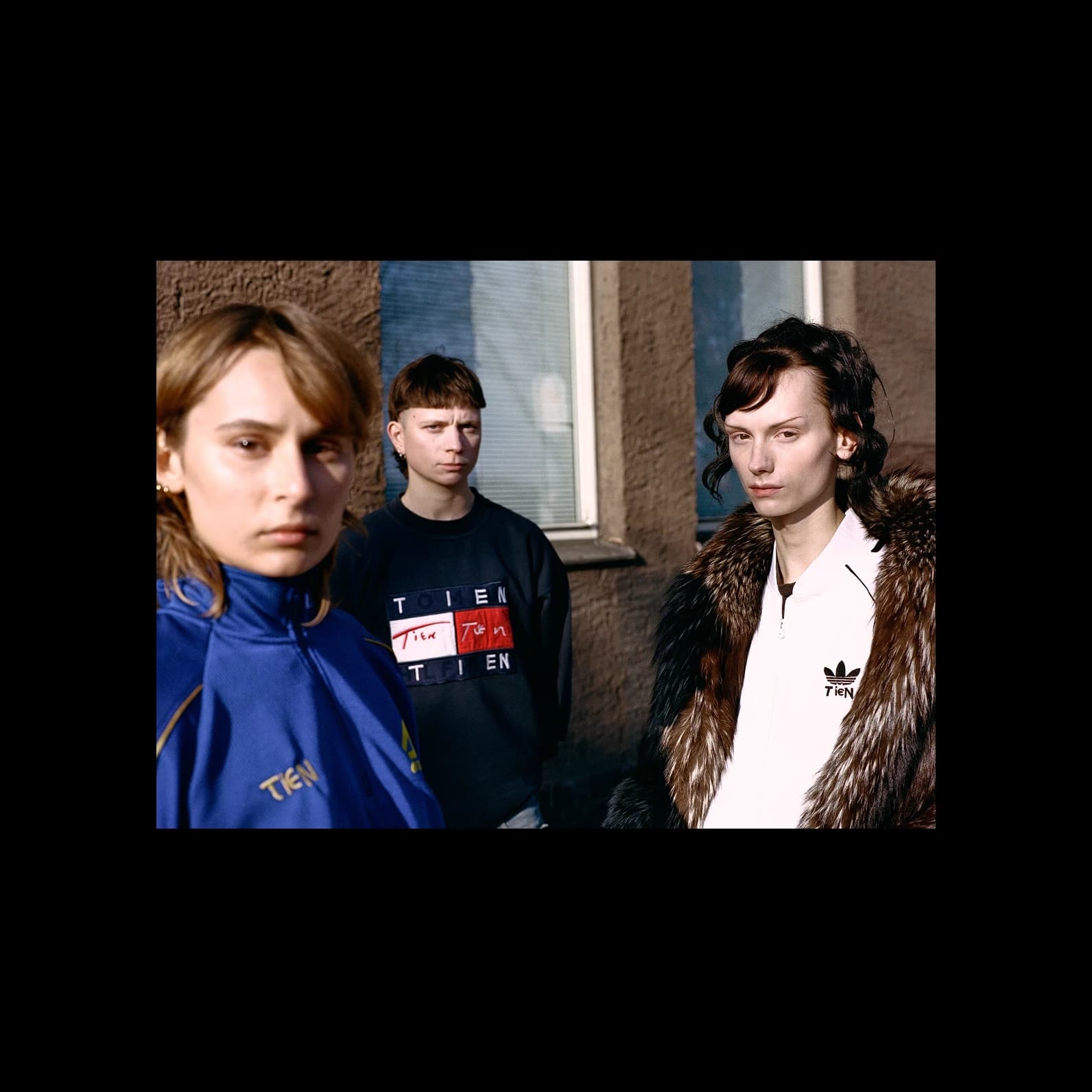 Three people wearing vintage sportswear reworked with the TIEN identity: an Adidas track jacket with TIEN embroidered on the chest, the Tommy Hilfiger sweatshirt with the flag logo replaced, and an Adidas zip-up with TIEN on the left chest.