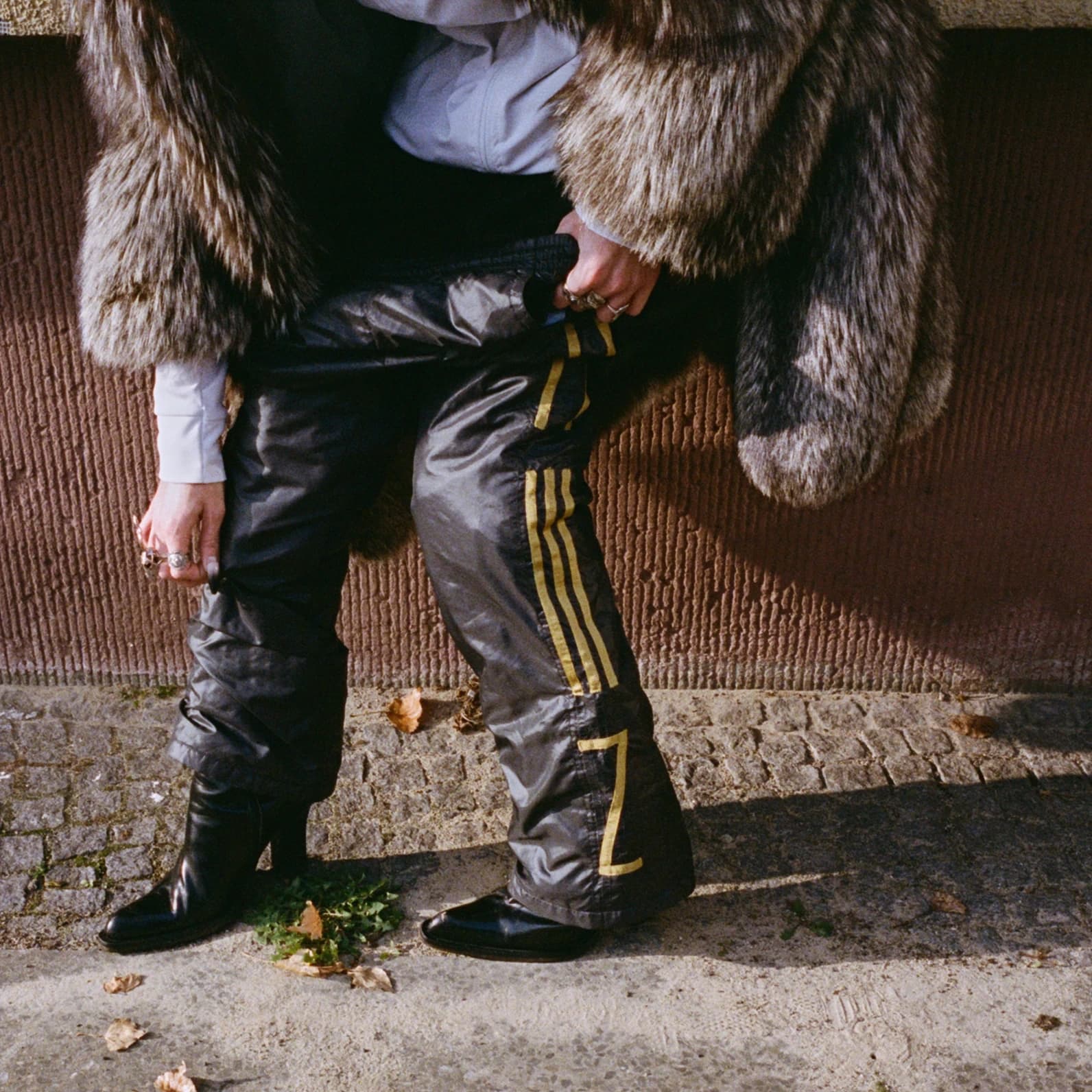 Close-up of a person standing on a cobblestone street wearing leather boots, a fur coat and black trackpants with yellow side stripes, shot for the TIEN campaign.