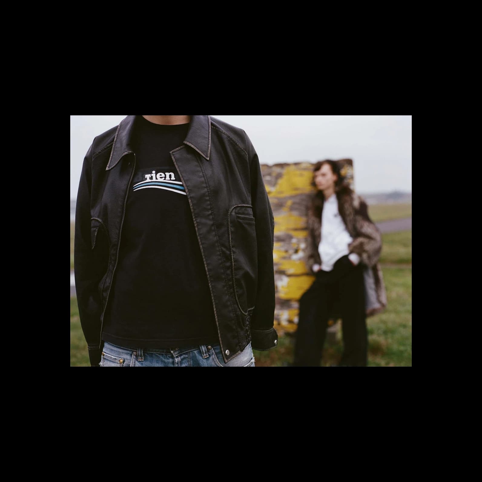 Person wearing a black t-shirt with a vintage Patagonia logo reworked to read "tien", layered under a black leather jacket, with a second person out of focus in the background.