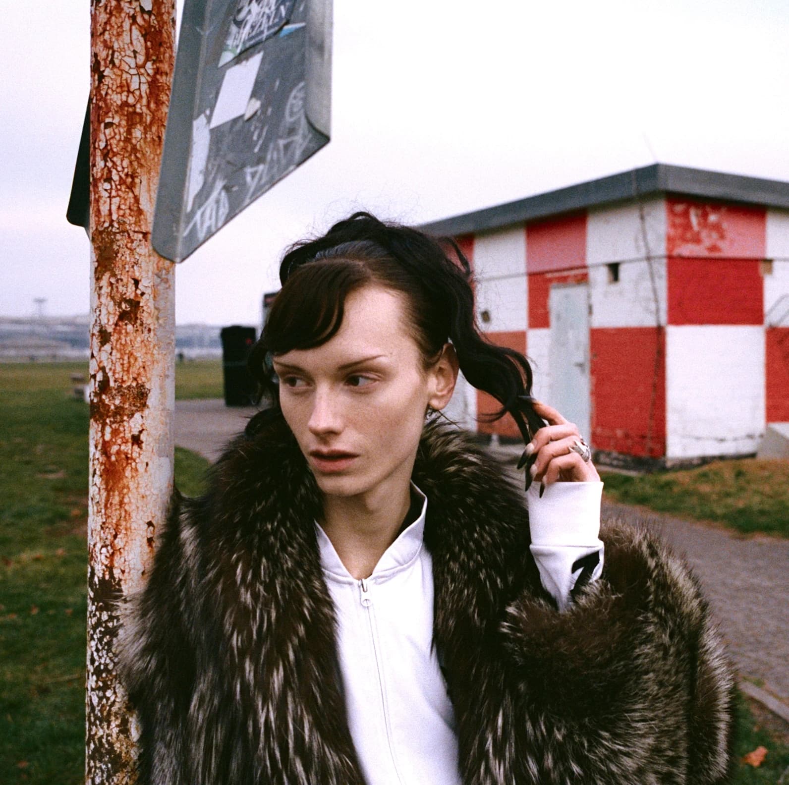 Person wearing a fur coat over a white Adidas zip-up with TIEN on the chest, standing next to a rusted street pole with a red and white checkered building in the background.