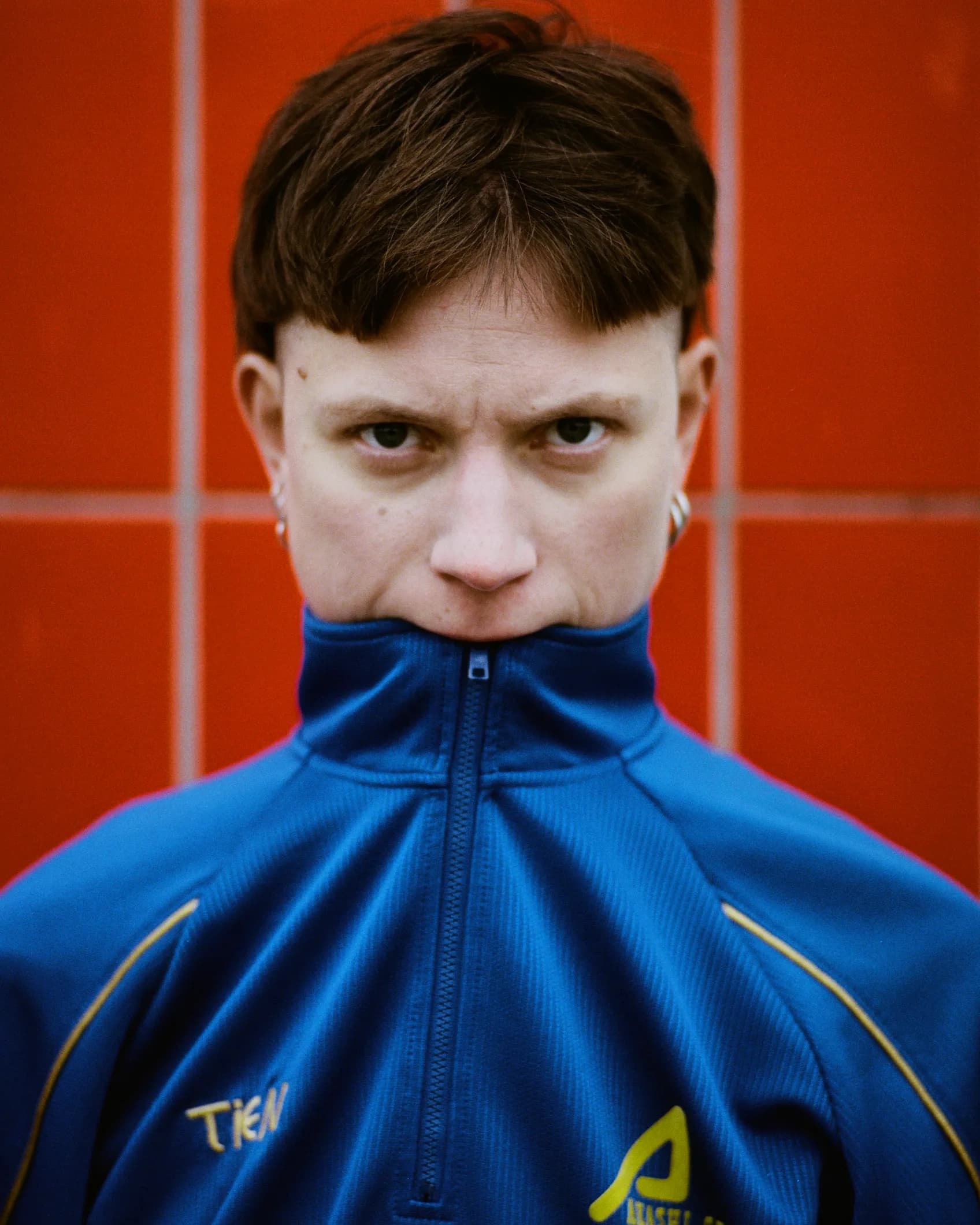 Close-up portrait of a person with a blue zip-up track jacket pulled up to their nose, shot against a red tiled wall for the TIEN campaign.
