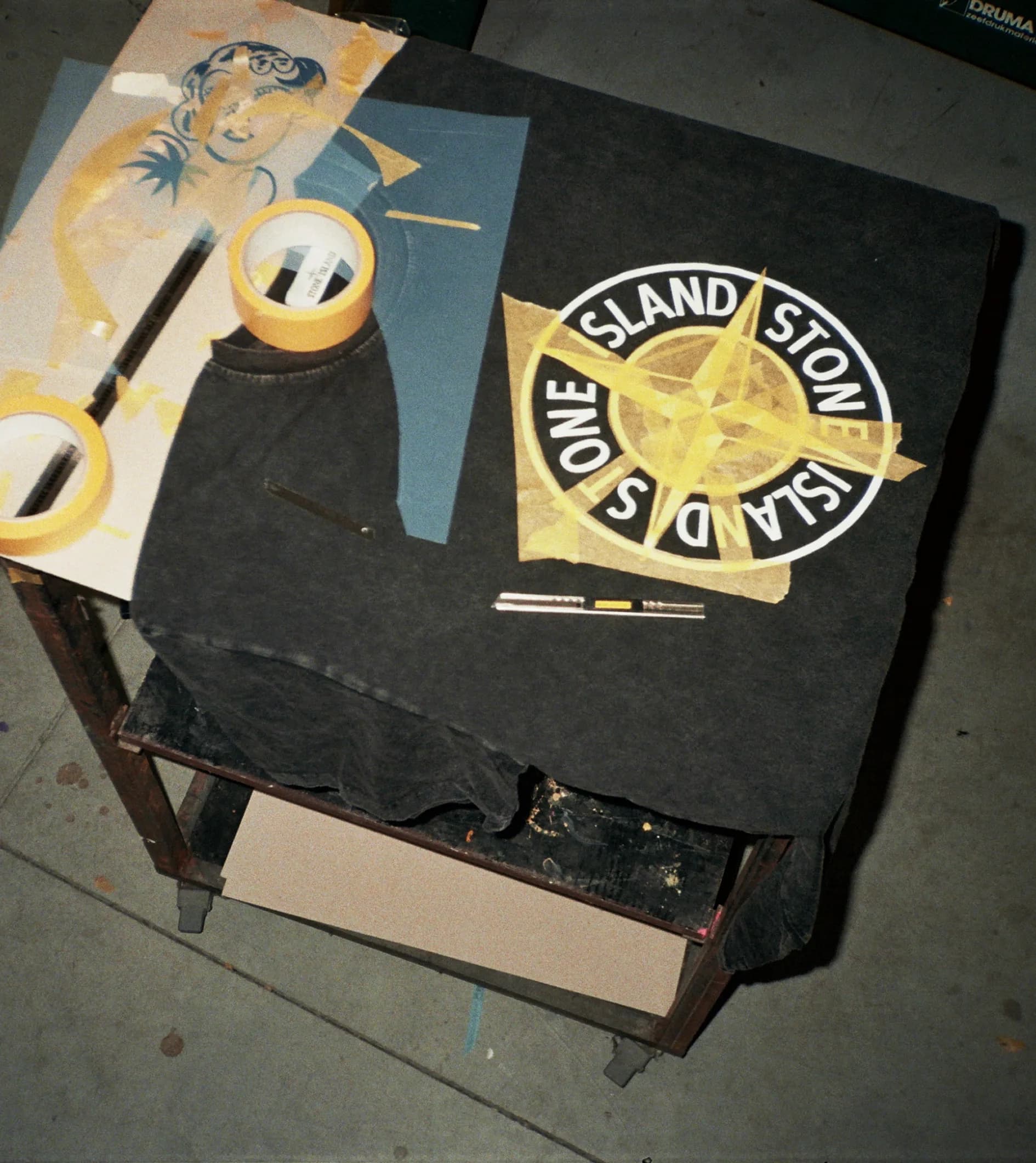 Behind the scenes shot of a Stone Island t-shirt laid flat on a work table with masking tape and acetate stencil in place, mid-process of being reworked with the TIEN identity.