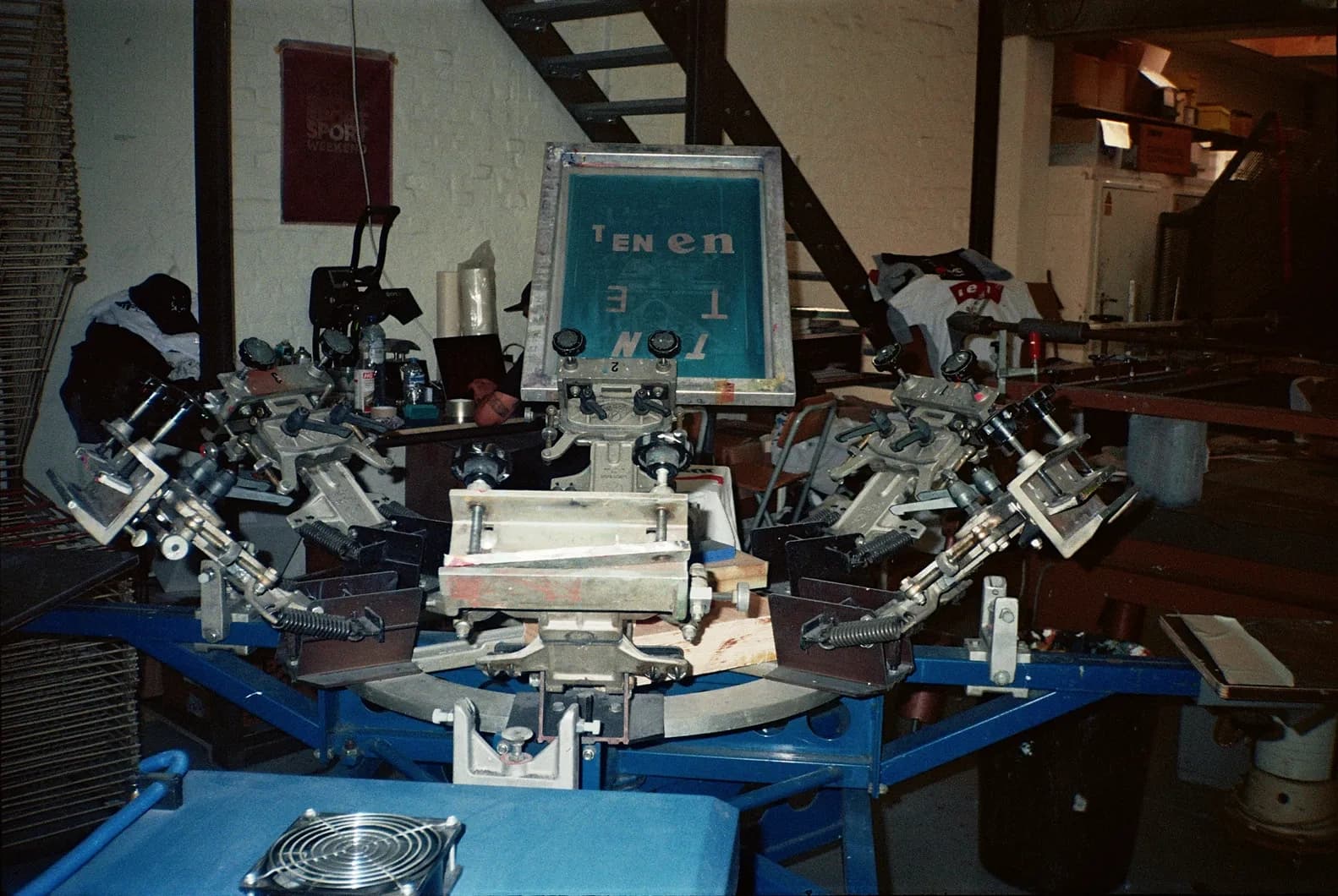 Behind the scenes shot of a multi-station rotary silkscreen press with a TIEN screen mounted, surrounded by finished and unfinished garments in the print shop.