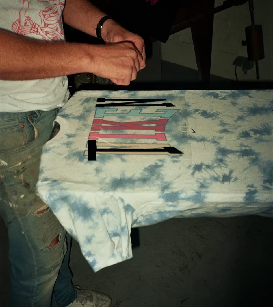 Behind the scenes close-up of the tie-dye Nike t-shirt mid-print, with the TIEN varsity wordmark being silkscreened onto the chest in layers of teal, pink and black.